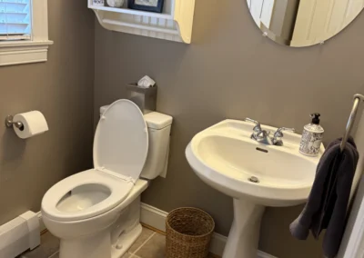 A cozy bathroom featuring a white toilet, sink, and decorative shelf with framed art, towels, and a woven basket on tiled flooring