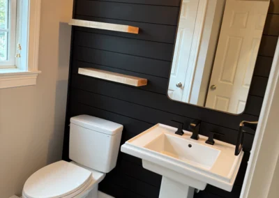 A modern bathroom featuring a black shiplap wall, a white pedestal sink, a toilet, and two wooden shelves illuminated by stylish wall sconces