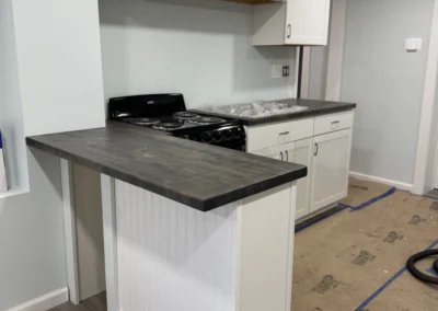 A modern kitchen with white cabinets, a black stove, and a wooden countertop is partially completed, surrounded by remodeling materials