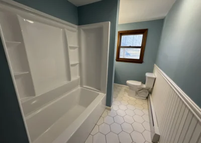 A bright bathroom featuring blue walls, a white hexagonal tile floor, a sink with a wooden cabinet, and a tub with a showerhead.