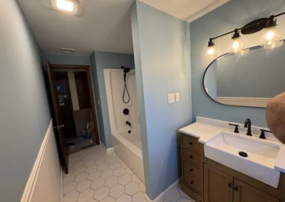 A bright bathroom featuring blue walls, a white hexagonal tile floor, a sink with a wooden cabinet, and a tub with a showerhead.