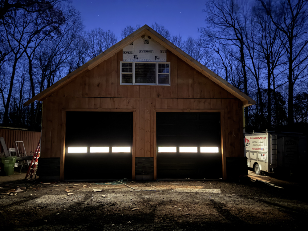 Garage Builds A newly constructed wooden garage at dusk, featuring illuminated garage doors and surrounding trees silhouetted against a dark blue sky.