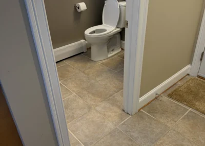 View of a light-colored bathroom with a white toilet, tile flooring, and a small shelf decorated with towels and a framed quote