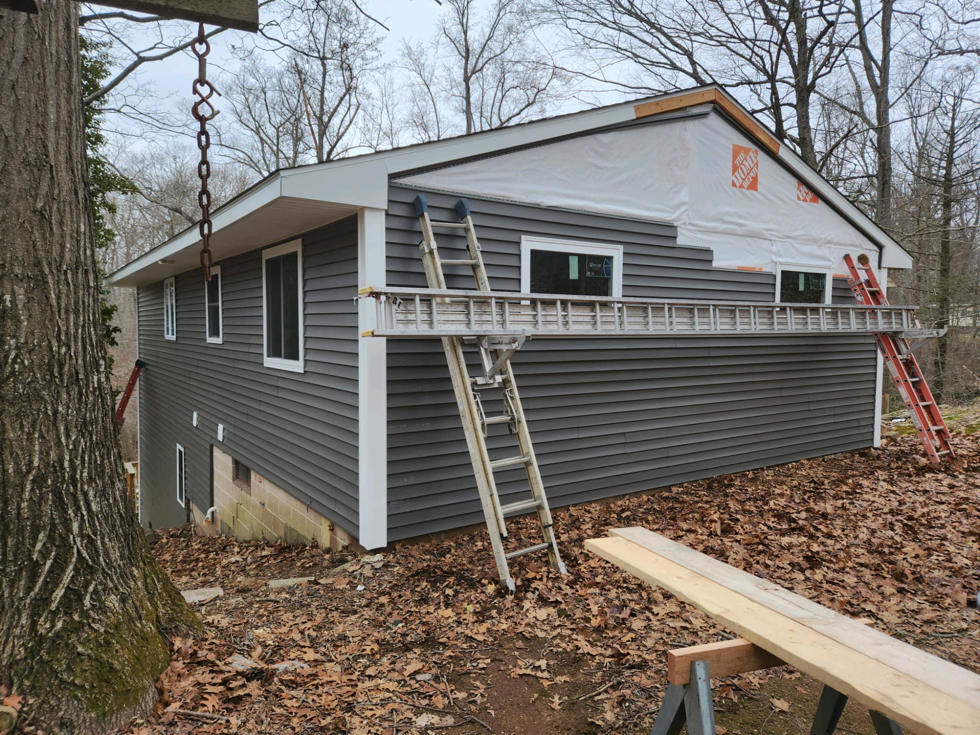 House on a sloped lot undergoing exterior renovation with new dark gray vinyl siding being installed. The upper portion is still wrapped in Home Depot-branded house wrap. Several ladders and construction tools are positioned around the site, with fallen leaves covering the ground. Located in Andover, Connecticut.